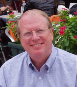 A white, red-haired man with glasses and a light blue shirt. Flowers in the background