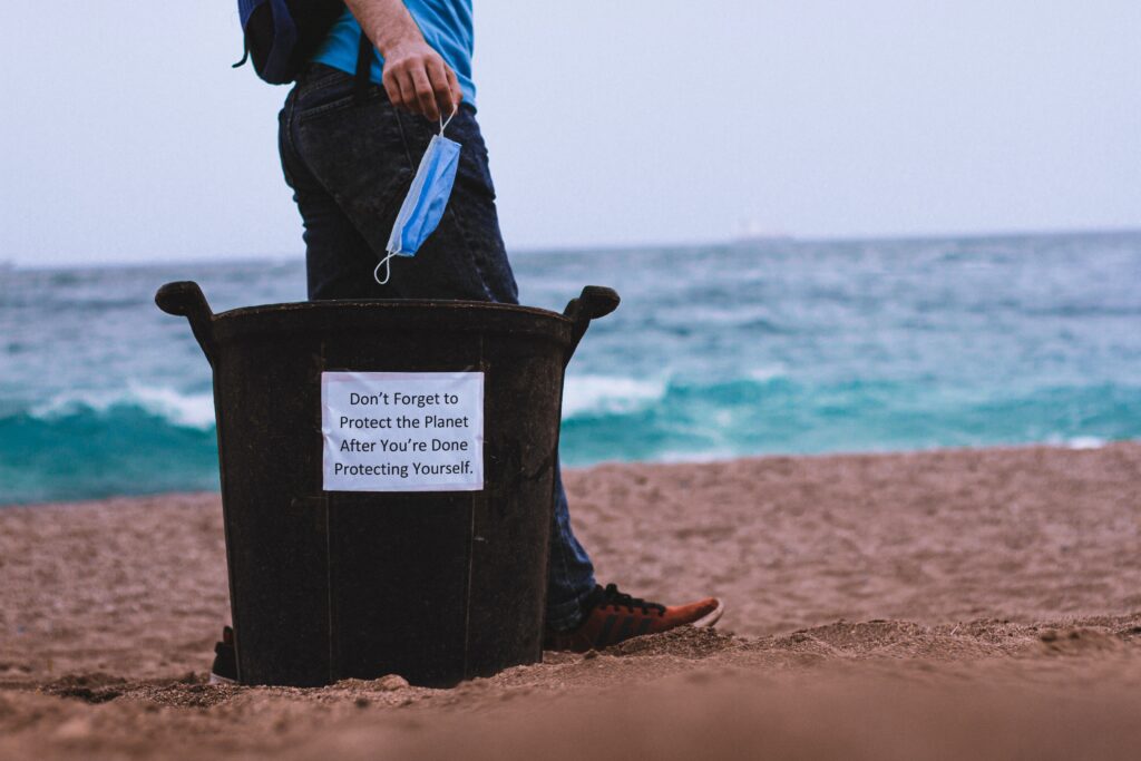 Person dropping a face mask into a trash can on the beach.