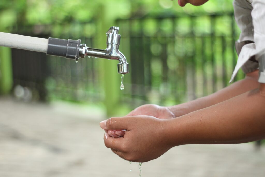 Person cupping their hands below a faucet dripping water. A fence and greenery in the background